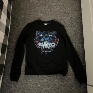 Svart Kenzo tröja med tigertryck - Säljer en snygg svart tröja från Kenzo med ett coolt tigertryck i blått och rött på framsidan. Tröjan har långa ärmar och en normal passform. 