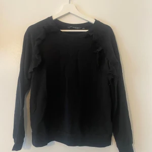 Svart tröja med volangdetaljer från NOSH - Säljer en stilren svart tröja från NOSH i storlek M. Tröjan har långa ärmar och snygga volangdetaljer längs axlarna som ger en extra touch. Perfekt för både vardag och fest! 🖤