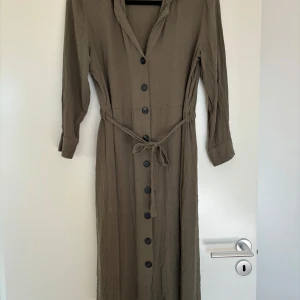 Klänning  - Khakigrön/olivgrön klänning med knappar och knyte från märket Pigalle i strl. 38. Sparsamt använd och i fint skick. 
