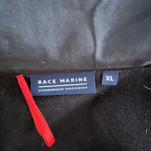 Svart jacka från Race Marine - Säljer en svart jacka från Race Marine i storlek XL. Den har en dragkedja framtill och en huva för extra skydd mot väder och vind. Perfekt för höst- och vårsäsongen. Jackan har en stilren design med Race Marine-loggan på ärmen.