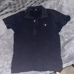 Svart pikétröja från Gant - Snygg svart pikétröja från Gant med klassisk krage och knappdetalj framtill. Perfekt för en stilren och avslappnad look. Väl använd