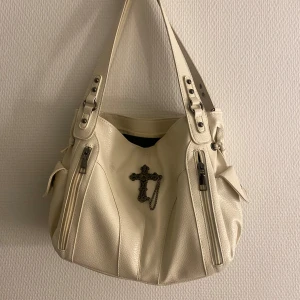 Vit axelväska med korsdetalj - Snygg vit axelväska i syntetmaterial med coola dragkedjor och en unik korsdetalj på framsidan. Väskan har justerbara remmar med nitar och praktiska sidofickor med knappar. Perfekt för att ge din outfit en edgy touch!