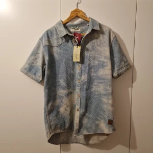 Blå kortärmad skjorta från Anerkjendt XL - Snygg kortärmad skjorta från Anerkjendt i en cool jeansblå färg. Skjortan har ett unikt mönster och knappar framtill. Perfekt för en avslappnad stil med en touch av dansk design.