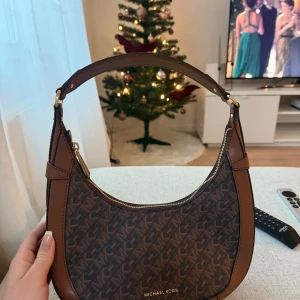 Brun axelväska från Michael Kors - Snygg brun axelväska från Michael Kors med ett diskret mönster och guldfärgade detaljer. Köpt i Paris i somras men har tyvärr inte kommit till användning, helt i nyskick💞köpt för ca 3000kr 