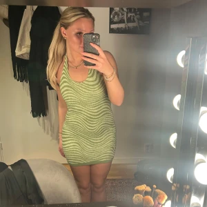 Grön randig klänning - Snygg grön klänning med randigt mönster och halterneck-design. Den är tight och kort, perfekt för stranden. Klänningen har en öppen rygg som ger en extra touch av stil.