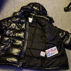 Svart moncler maya  - Säljer en snygg svart pufferjacka från Moncler en gamlare jacka.Jackan har en glansig finish och är utrustad med en dragkedja framtill. Den har en broderad Moncler-logga på ärmen och en praktisk huva för extra skydd mot väder. Perfekt för kyligare dagar! 