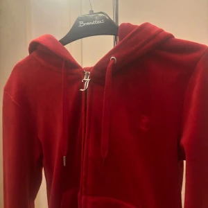 Röd hoodie från Juicy Couture - Snygg röd hoodie från Juicy Couture i mjukt material. Den har en dragkedja framtill och en praktisk huva med snören. Perfekt för en avslappnad stil.