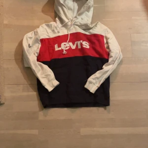 Flerfärgad hoodie från Levi's - Säljer en snygg flerfärgad hoodie från Levi's i vitt, rött och blått. Tröjan har en klassisk design med en stor logga på bröstet och en bekväm huva med dragsko. Storlek S men kan passa M