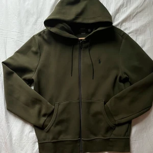 Olivgrön hoodie från Polo Ralph Lauren - Säljer en stilren olivgrön hoodie från Polo Ralph Lauren med dragkedja och broderad logotyp på bröstet. Tröjan har långa ärmar och en praktisk huva med dragsko. Nypris 2.295:- SEK