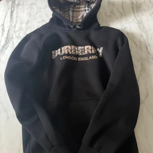Svart hoodie från Burberry - Snygg svart hoodie från Burberry med klassisk logga på bröstet och rutigt mönster i luvan. Perfekt för en stilren och bekväm look. Den har långa ärmar och en praktisk magficka framtill.