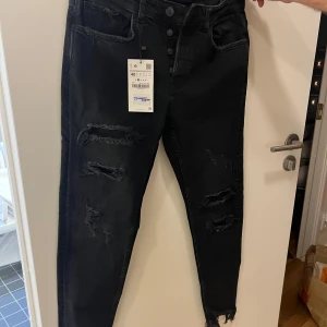 Svarta jeans med slitningar - Snygga svarta jeans från Zara med slitningar på framsidan. De har en hög midja och knappar framtill. Perfekta för en trendig look med en skinny passform.