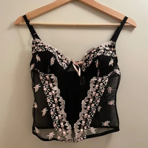 Svart korsett med blommiga detaljer  - Jättefin svart korsett med rosa och vit blommiga detaljer. Den har justerbara axelband och en söt rosett framtill. Aldrig använt denna då den är för liten, märker står tyvärr inte men det är mycket bra skick. Storleken är 75b.