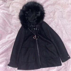 Svart kappa med fauxpäls - Svart kappa med fauxpäls runtom luvan. Små defekter på läderdetaljerna och en liten ljus fläck på fickan(se bilder). Använd gärna köp nu💗