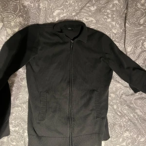 Grön cardigan - Säljer en stilren grön cardigan med dragkedja och långa ärmar. Tröjan har en klassisk krage och två fickor framtill. Perfekt för en avslappnad look.