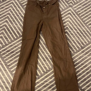 Bruna byxor med utsvängda ben - Snygga bruna byxor med hög midja och utsvängda ben. Perfekta för en trendig look. Jeansen  har en dragkedja framtill och är tillverkade i ett mjukt material.  Storlek 26