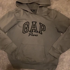 Grå hoodie från GAP - Säljer en grå hoodie från GAP med texten 'GAP Paris' på framsidan. Tröjan har en klassisk känguruficka och långa ärmar. Perfekt för en avslappnad stil. Litet hål på armen så priset kan diskuteras, sitter mer som s eller xs men är m
