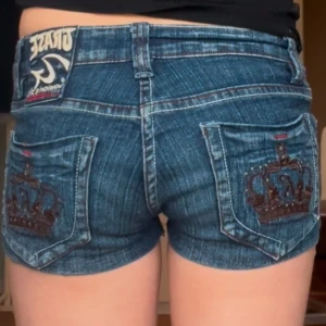 Jeans shorts - Snygga blå jeansshorts från Grace med broderade kronor på bakfickorna. De har en låg midja och en dragkedja framtill. Perfekta för en avslappnad stil. Tyvärr så har knappen gått av men det märks inte om man spänner fast med en säkerhetsnål!❤️