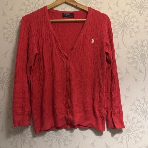 Röd kofta från Polo Ralph Lauren - Säljer en snygg röd kofta från Polo Ralph Lauren med kabelstickat mönster och broderad logga på bröstet. Koftan har en v-ringad design och knappar framtill. Perfekt för en stilren look!