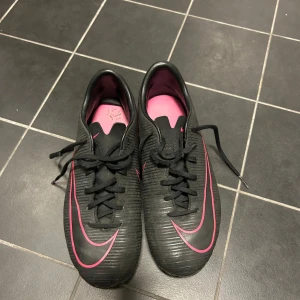 Svarta fotbollsskor från Nike - Säljer ett par svarta Nike fotbollsskor med rosa detaljer och snörning. Skorna har en sportig design med dobbar för bra grepp på planen. Perfekta för fotbollsträning och matcher.