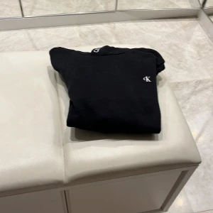 Svart hoodie från Calvin Klein - Säljer en stilren svart hoodie från Calvin Klein med deras logga diskret på bröstet. Hoodien har långa ärmar och en bekväm huva med Calvin Klein Jeans-tryck på insidan. Perfekt för en avslappnad look.