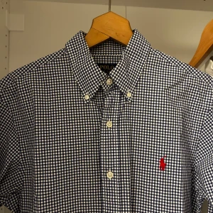 Rutig skjorta från Ralph Lauren - Snygg rutig skjorta från Ralph Lauren i blått och vitt. Skjortan har en klassisk krage med knappar och en broderad logotyp på bröstet. Perfekt för en stilren look.