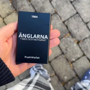 Korthållare ÄNGLARNA - PushWallet UF  - Säljer en stilren svart PushWallet med texten 'Änglarna - I med och motgång' och årtalet 1904. Perfekt för att hålla dina kort säkra och organiserade. Kompakt och lätt att bära med sig.