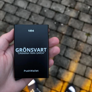 Korthållare GRÖNSVART - PushWallet UF  - Säljer en stilren svart PushWallet plånbok med texten 'GRÖNSVART DARARNA FRÅN GÅRDA' och årtalet 1894 tryckt på framsidan. Perfekt för att hålla dina kort säkra och organiserade. Enkel och smidig design som passar i fickan. Helt ny.
