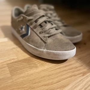 Beiga sneakers från Converse - Snygga beiga sneakers från Converse i mocka med klassisk stjärndetalj i blått på sidan. Skorna har en vit sula och snörning framtill. Perfekta för en avslappnad stil.