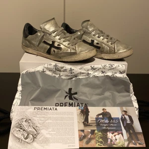 Silvriga sneakers från Premiata - Snygga silvriga sneakers från Premiata White Edition med svarta detaljer. Skorna har en unik design med en sliten look och är perfekta för en trendig stil. Säljer dem på grund av att de börja bli för små. Ny pris 2999kr jag säljer för 429. Priset år på grund av att de är ganska slitna men har lite kvar och ge. Tror att de är allt og som följer med allt syns på bilderna. Priset kan diskuteras vid snabb affär. Hör av er om ni har frågor! MVH Theodor!