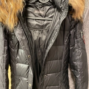 Svart dunjacka med päls från Rockandblue - Snygg svart dunjacka från Rockandblue med en lyxig pälsdetalj på huvan. Jackan har en quiltad design och dragkedja framtill. Perfekt för kalla dagar med sin långa passform och stilrena look.