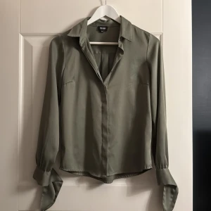 Olivgrön skjorta  - 100% polyester, känns dock som silke. Säljer pga. för liten idag.  En maska på andra bilden, i övrigt fint skick.  Knyts i ärmarna