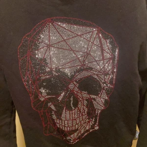 Svart tröja med dödskalle från Philipp Plein - Snygg svart tröja från Philipp Plein med en stor, glittrig dödskalle på framsidan. Tröjan har långa ärmar och en rund halsringning. Perfekt för en cool och edgy stil.