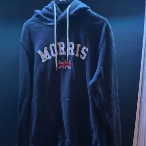 Morris Hoodie  - Säljer en snygg mörkblå hoodie från Morris med broderad text och brittisk flagga på framsidan. Tröjan har en klassisk design med dragsko i huvan och långa ärmar. Perfekt för en avslappnad stil. Storlek Large men passar även Medium!   Tveka inte att höra av er vid funderingar🥂