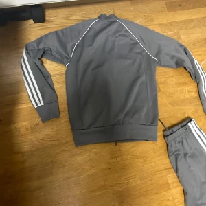 Grå träningsdräkt från Adidas SST - Säljer en grå träningsdräkt från Adidas med klassiska vita ränder längs ärmar och ben. Jackan har dragkedja och byxorna har elastisk midja. Perfekt för träning eller en avslappnad dag.