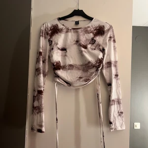 Mönstrad långärmad topp - Säljer en unik långärmad topp med ett abstrakt mönster i bruna och vita toner. Toppen har snörning i sidorna för en justerbar passform. Perfekt för att ge din outfit en konstnärlig touch.