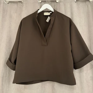 Neo noir blus - Snygg brun blus från Neo Noir med trekvartsärm och en stilren v-ringad design. Perfekt för en chic och avslappnad look. Blusen har en lös passform som ger en bekväm känsla.