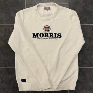 Vit stickad tröja från Morris - Säljer en stilren vit stickad tröja från Morris med ett tryck av Union Jack och texten 'Morris Great Britain Tradition' på framsidan. Tröjan har en rund halsringning och ribbade muddar vid ärmslut och nederkant. Perfekt för en klassisk look.