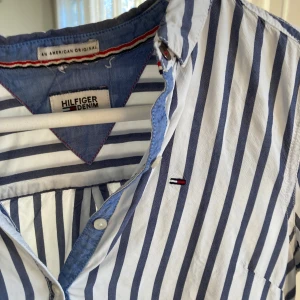 Randig skjorta från Hilfiger Denim - Snygg vit och blå randig skjorta från Hilfiger Denim. Skjortan har långa ärmar och knappar framtill. Perfekt för en stilren look med en touch av casual. Den har en klassisk krage och ett litet broderat märke på bröstet.