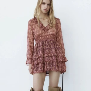 Mönstrad kortklänning med volanger - Trendig kortklänning med ett unikt mönster i varma toner. Klänningen har långa ärmar och flera volanglager som ger en lekfull look. Perfekt för att sticka ut med stil. Liten i storleken så passar S/XS, använd gärna KÖP NU