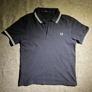 Svart pikétröja från Fred Perry - Snygg svart pikétröja från Fred Perry med vita detaljer på krage och ärmslut. Tillverkad i 100% bomull för en bekväm passform. Perfekt för en stilren look.