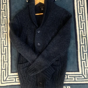 Mörkblå stickad cardigan - Säljer en mysig mörkblå stickad kofta med stora knappar och ribbade detaljer. Perfekt för kyliga dagar. Koftan har långa ärmar och två praktiska fickor framtill. Hör av vid fler frågor!