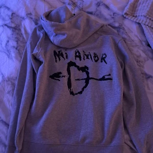 Grå hoodie från Mira Paris - Säljer en grå hoodie från Mira Paris med texten 'Mi Amor' och ett pilmotiv på ryggen. Tröjan har en klassisk design med långa ärmar och en bekväm huva. Perfekt för en avslappnad stil. Finns en liten defekt som är svart vid höger sida arm.