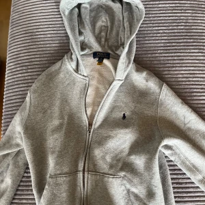 Grå hoodie från Polo Ralph Lauren - Säljer en snygg och bekväm grå hoodie från Polo Ralph Lauren. Den har en dragkedja framtill och en liten broderad logga på bröstet. Perfekt för höst och vinter, och passar både till vardags och chill. Den är i bra skick och väldigt mysig att ha på sig. Köp den nu eller ge ett bud! 💙