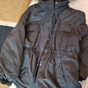 Svart dunjacka från Tommy Jeans - Säljer en svart dunjacka från Tommy Jeans i storlek L. Jackan har en mysig pälskant på huvan och är perfekt för kalla vinterdagar. Den har flera praktiska fickor och en dragkedja framtill. Jackan är i bra skick och håller dig varm och bekväm hela vintern. Passar perfekt för både vardag och utomhusaktiviteter.