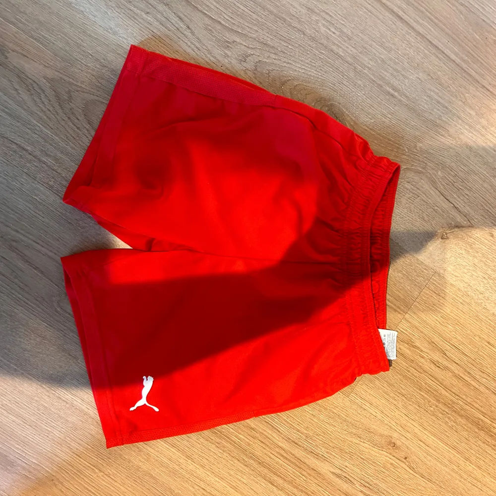 Säljer ett par röda träningsshorts från Puma. De är i nyskick och har en elastisk midja för en bekväm passform. Perfekta för träning eller fotboll. Puma-loggan är vit och sitter på ena benet. Sköna och lätta att röra sig i!. Shorts.