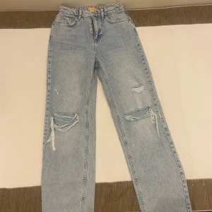 Ljusa jeans med slitningar - Säljer ett par ljusa jeans med slitningar på knäna och låren. De är i en straight fit och har en hög midja. Perfekta för en avslappnad och trendig look. Passar bra till både vardag och fest!