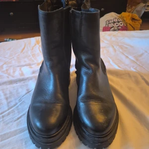 Svarta boots från Dasia - Säljer ett par svarta boots från Dasia i skinn. De har en dragkedja på sidan och elastiska paneler för enkel på- och avtagning. Skorna har en robust sula som ger bra grepp och är perfekta för höst och vinter. De är i bra skick och redo för en ny ägare!