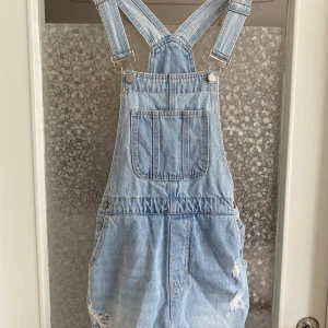 Jeansbyxdress med hängslen - Säljer en snygg jeansbyxdress med hängslen. Den är ljusblå och har en stor ficka på bröstet samt två fickor på sidorna. Byxdressen har en avslappnad passform och är perfekt för en casual look. Den har också slitna detaljer som ger en cool och trendig känsla.