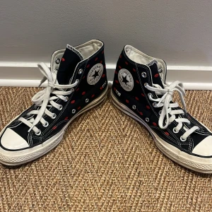 Svarta Converse Chuck Taylor med röda detaljer - Säljer ett par svarta Converse Chuck Taylor sneakers med vita snören och röda pussmunnar. Skorna har en klassisk hög modell med vit sula och tåhätta. De är bara använda en gång alltså är dem i nyskick. Dem är från en allahjärtansdags kollektion och såldes då för 90$(sista bilden).Perfekta för en casual look!💋
