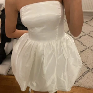 Vit axelbandslös klänning - Säljer en superfin vit axelbandslös klänning. Den är kort och har en fluffig kjol som ger en söt och festlig look. Perfekt för sommarfester eller studentfirande! Klänningen är i nyskick och har en tight passform upptill som framhäver figuren.